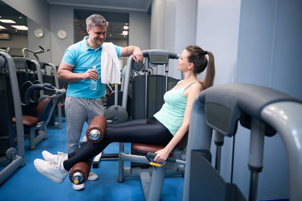 Smiling female exercising on leg extension machine while talking to pleased training companion - Photo, Image