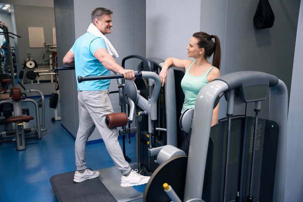 Pleased sporty man performing side leg raise on exercise machine in presence of smiling fitness partner - Photo, Image