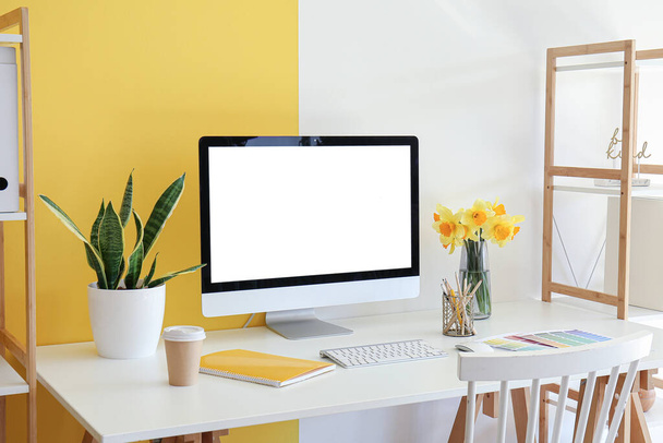 Современный компьютер, красивые нарциссы цветы, кофеварка, ноутбук и краски цветовой палитры на столе рядом с цветной стеной - Фото, изображение