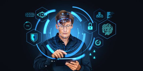 Serious young European businessman in blue shirt using tablet computer working with facial recognition interface over dark blue background. Concept of biometric scanning - Φωτογραφία, εικόνα