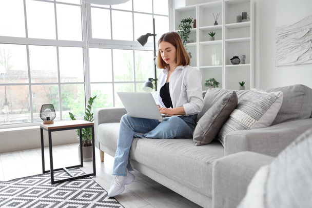 Pretty young woman sitting on grey sofa and using modern laptop in light living room - Photo, Image