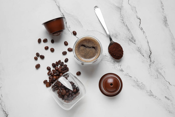 Xícara de café expresso delicioso, vidro virado com feijão, cápsulas e colher na mesa branca - Foto, Imagem