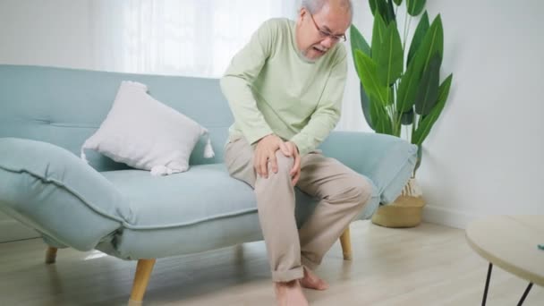 Старший мужчина с помощью руки трогать колено в точке боли, пожилой человек страдает от боли в колене, сидя на диване дома, люди с концепцией проблем со здоровьем - Кадры, видео