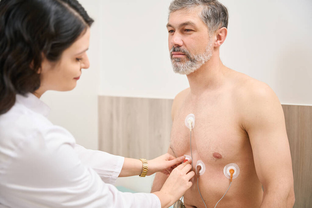 Кардиолог в белом халате готовит пациента к электрокардиографии сердца, мужчина смотрит на врача - Фото, изображение
