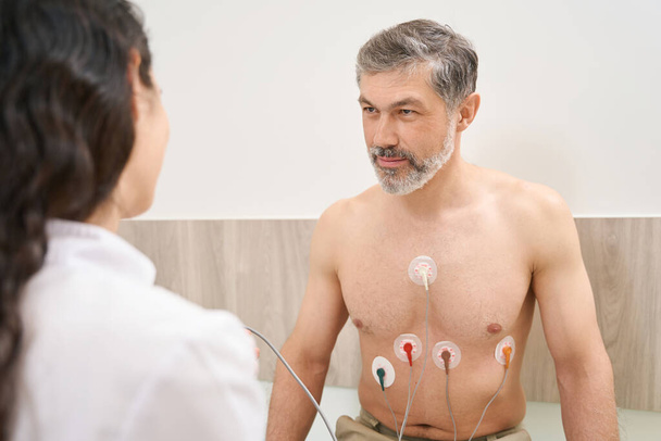 Мужчина сидит рядом с медсестрой и проходит электрокардиографию сердца в больнице - Фото, изображение