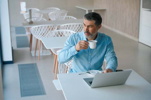 Взрослый мужчина сидит на стуле за столом и пьет чай, печатает на ноутбуке в столовой - Фото, изображение
