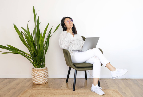 Pensativo joven mujer asiática que trabaja en el ordenador portátil en sillón en el interior. Espacio para texto. - Foto, Imagen