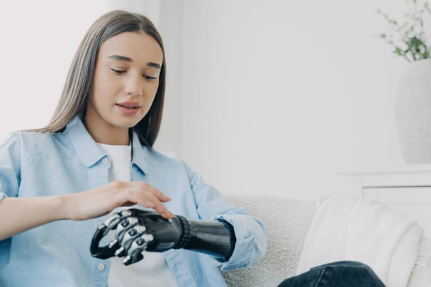 Handicapped girl explores bionic arm, presses buttons. Young European woman with cyber hand at home. Futuristic bionic prosthesis tech. - Photo, Image