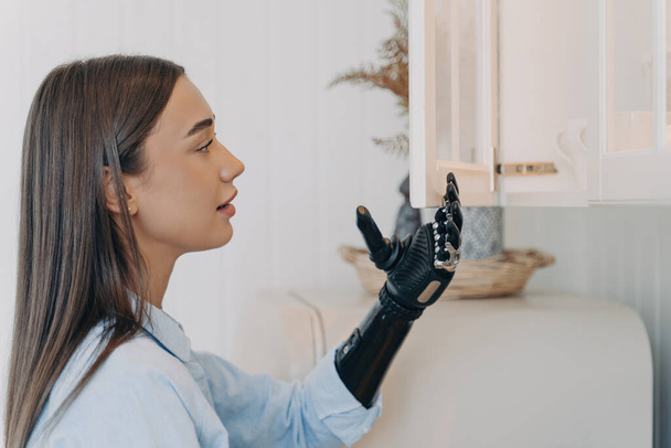 Handicapped girl opens cupboard with cyber hand. European woman with artificial arm. Modern bionic prosthesis functions. Disabled person's routine. - Photo, Image