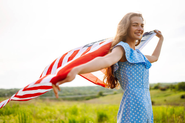 Young woman with american flag on blooming meadow. 4th of July. Independence Day. Patriotic holiday. USA flag fluttering in the wind. - Φωτογραφία, εικόνα