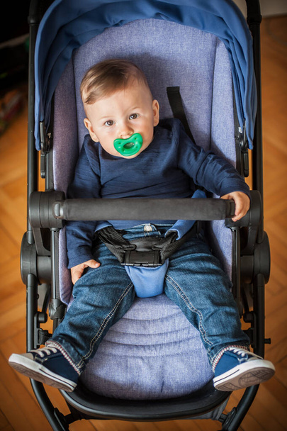 Portrait of cute baby boy in jeans and shoes, in pram baby carriage, looking at camera. Love and family emotion - Zdjęcie, obraz
