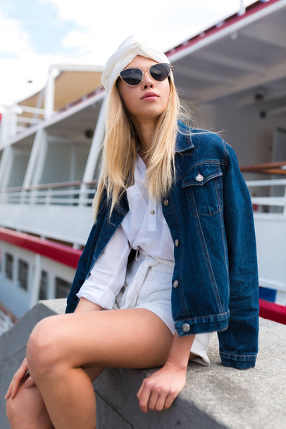 Улыбающаяся модная женщина в модных солнцезащитных очках, сидящая против лодки в городе. Красивая летняя женщина в джинсовой куртке отдыхает на открытом воздухе и весело проводит выходные - Фото, изображение
