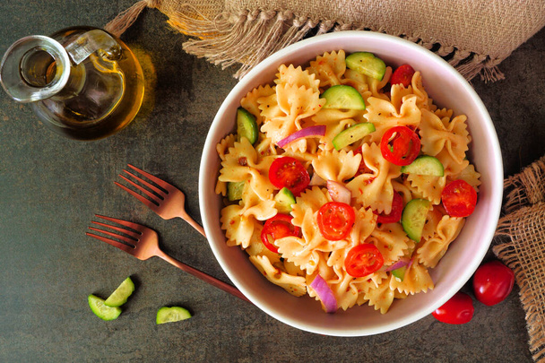 Traditional pasta salad with tomatoes, onion and cucumbers. Top down table scene on a dark stone background. - Photo, Image