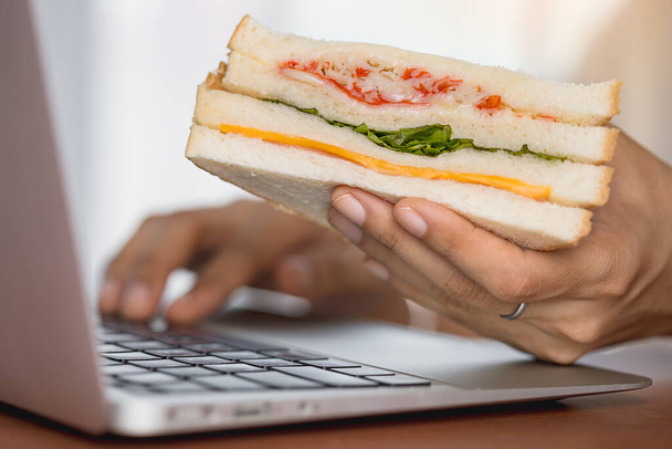 Kädessä pitämällä herkullinen voileipä ja käyttämällä kannettavaa tietokonetta syömiseen työskennellessään käsite. - Valokuva, kuva