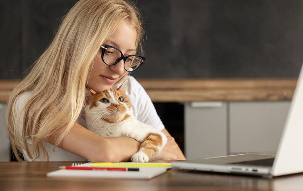 ラップトップを使用している若い女性とかわいい猫がキーボードに座っています。カジュアルな女の子仕事上のラップトップとともに彼女のペット中に座っている家に居心地の良いオフィス. - 写真・画像