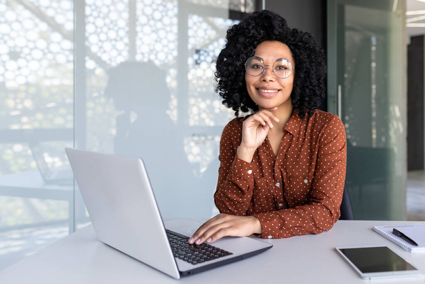Porträt einer erwachsenen erfolgreichen Geschäftsfrau, afrikanisch-amerikanische Frau am Arbeitsplatz lächelnd und in die Kamera blickend, selbstbewusste Chefin am Arbeitsplatz im Büro, mit Laptop. - Foto, Bild