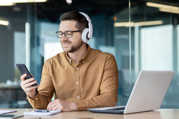A smiling young man in headphones and with a phone in his hands was sitting at a desk in the office. He works, rests, talks on a video call, listens to music. - 写真・画像