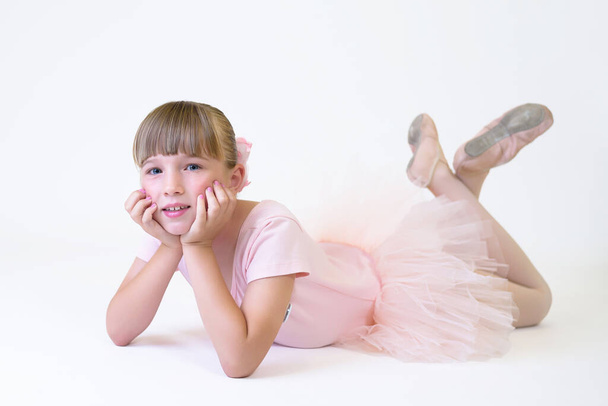 Mała tancerka baleriny w różowej tutu student akademii pozowanie na białym tle. Taniec dziewczyna leży na podłodze pozowanie do kamery. - Zdjęcie, obraz