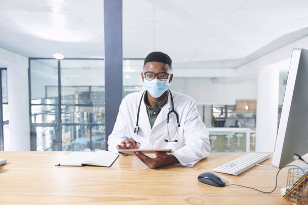 Llevo mi máscara para los dos. un joven médico con una máscara facial y sentado en su consultorio en la clínica mientras usa la tecnología - Foto, imagen