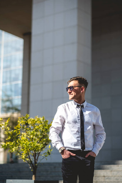 Jeune homme d'affaires professionnel, séduisant et confiant avec chemise blanche et cravate noire debout sous le soleil dans le parc urbain du quartier des affaires - Photo, image