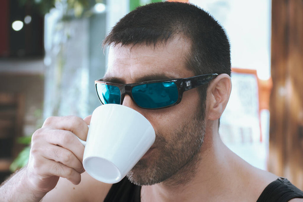 Modern man with sunglasses and sleeveless shirt savoring coffee up close, highlighting his fresh and urban style. An energy-filled and charming pause. - Фото, зображення