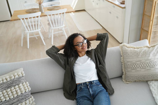 Boldog afro-amerikai lány pihen egy kényelmes kanapén egy hangulatos nappaliban, élvezi a jól megérdemelt szünetet. - Fotó, kép