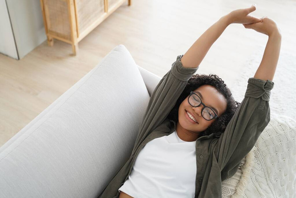 Happy mixed race girl stretches and relaxes on the sofa at home, enjoying a weekend afternoon nap for her wellness. - 写真・画像