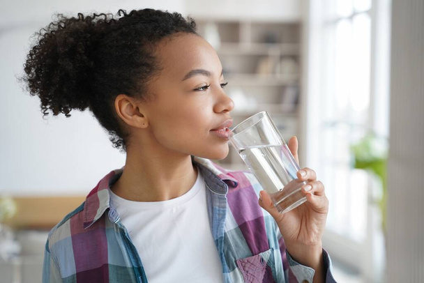 Chica sana de raza mixta disfruta de la rutina de la mañana, beber agua pura de vidrio en casa, abrazando un estilo de vida saludable. - Foto, imagen