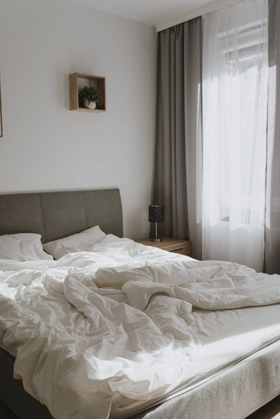 Aesthetic minimal home, bedroom interior design. Modern Nordic Scandinavian interior design. Bed with crumpled linens, pillow, window with curtains, sunlight shadows. Elegant cozy apartment for rent - Photo, image