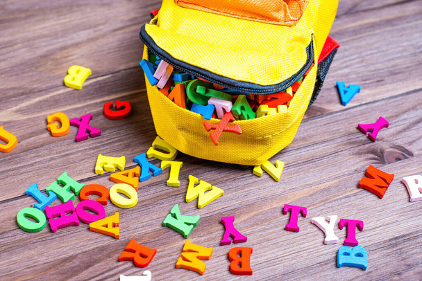 Small yellow and red backpack filled with wooden letters, playfully scattered on a wooden table. Vibrant preschool season concept. - Fotografie, Obrázek