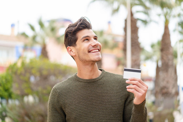 Young caucasian man holding a credit card at outdoors looking up while smiling - Photo, Image