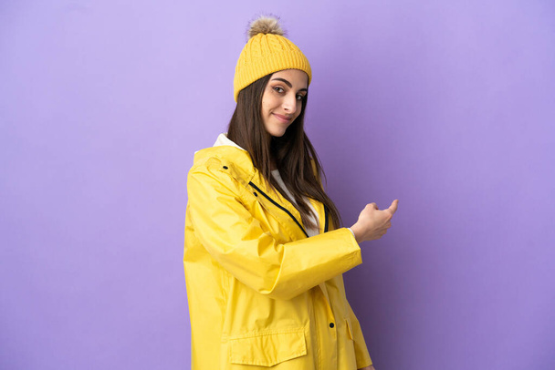 Young caucasian woman wearing a rainproof coat isolated on purple background pointing back - Foto, Imagem