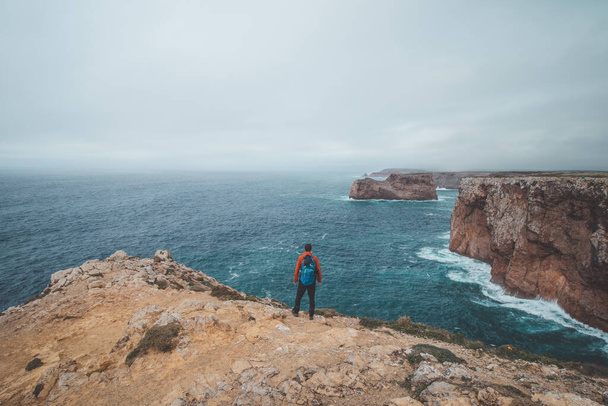 Der schwarzhaarige Abenteurer steht am Ende von Kap Cabo de Sao Vicente im Südwesten Portugals an der Algarve. Der Mensch genießt seine Freiheit. Wanderung auf dem Fischerpfad. - Foto, Bild