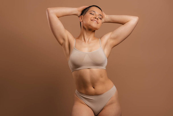 Happy young short hair woman in underwear holding hands behind head against grey background - Foto, imagen