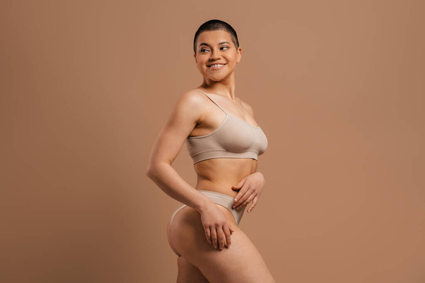 Confident young short hair woman in underwear smiling while standing against grey background - Foto, imagen