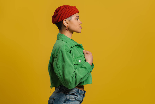 Side view of fashionable young woman in hat standing against yellow background - Foto, afbeelding