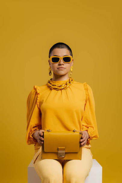 Stylish woman in fashionable glasses carrying trendy bag while sitting against yellow background - Fotografie, Obrázek