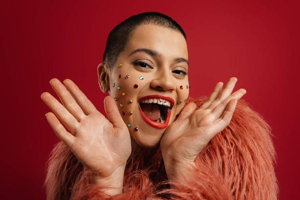 Surprised young short hair woman with shiny crystals over her face smiling against red background - Foto, Imagen