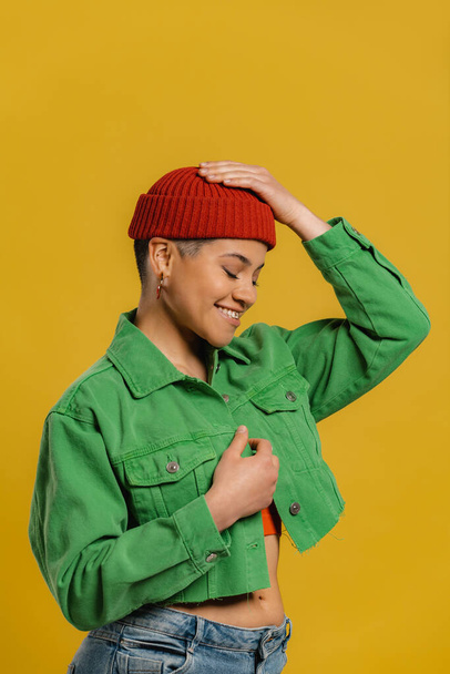 Fashionable young woman adjusting her hat and smiling while standing against yellow background - Foto, afbeelding