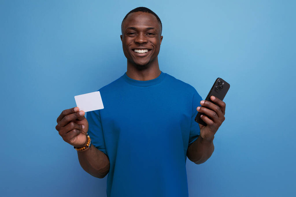 30-jähriger gutaussehender Mann mit dunkler Hautfarbe, bekleidet mit einem einfachen T-Shirt, das ein Smartphone und eine Plastikkarte für Einkäufe im Hintergrund mit Kopierplatz hält. - Foto, Bild