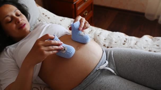 Bovenaanzicht van een zwangere vrouw die baby laarsjes op haar buik legt in het derde trimester van de late zwangerschap.Ze geniet van haar zwangerschapsstijl, ontspant zich thuis, ligt op het bed in de slaapkamer.Verwacht een baby - Video