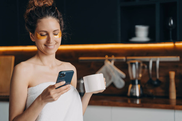 Young latina reads messages on smartphone. Happy girl with under-eye patches, towel-clad, chats, shops online in kitchen after morning shower at home. - Photo, Image