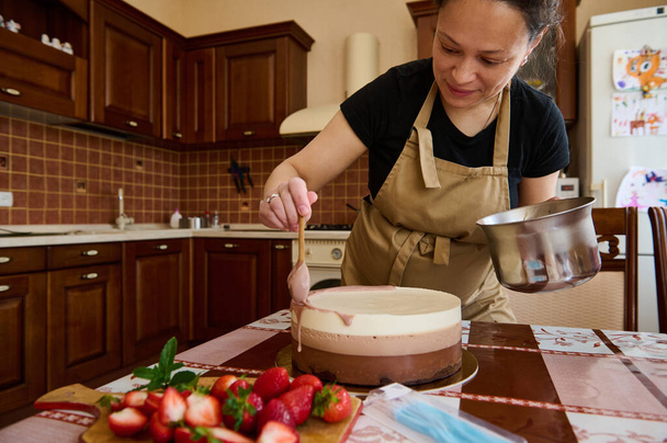 Πορτρέτο της εγκύου σεφ ζαχαροπλαστικής διακόσμηση με ρουμπινί ροζ γάνωμα της τούρτας γενεθλίων, στρώσεις με τρεις στρώσεις μους σοκολάτας, στην κουζίνα come. Ζαχαροπλαστική. Αρτοποιείο. Βιομηχανία τροφίμων - Φωτογραφία, εικόνα