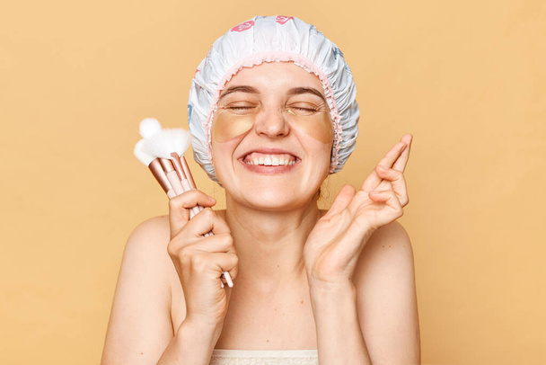 Extremely happy hopeful woman wearing shower cap holding makeup brushes keeps eyes closed crossing fingers making wish hopes for good luck stands isolated over beige background - Foto, afbeelding