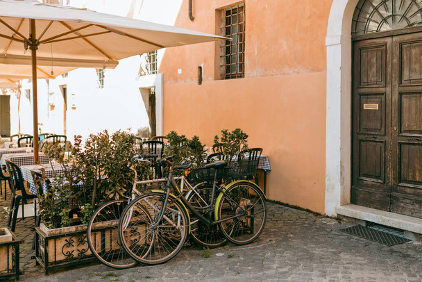 Bicycles parked on the street in Rome, Italy. Old bike against the orange wall at home. City transport concept. Lush green plants growing in pots near door of house - Photo, image