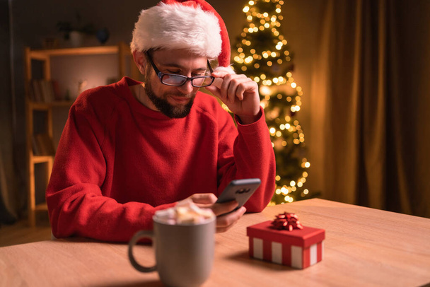 Caucasian man wearing a red sweater and glasses working late and using smartphone in a home office with a Christmas tree in the background, reading good news and smiling. Copy space - Photo, Image