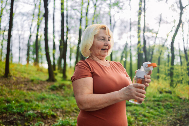 Close up portrait of an attractive senior 60s woman drinking water from bottle in the forest. High quality photo - Zdjęcie, obraz