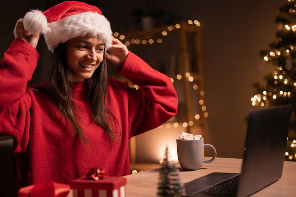 Ευτυχισμένη γυναίκα δείχνει ένα καπέλο Σάντα, ενώ έχουν βιντεοκλήση πάνω από το φορητό υπολογιστή την παραμονή των Χριστουγέννων στο σπίτι. Αντιγραφή χώρου - Φωτογραφία, εικόνα