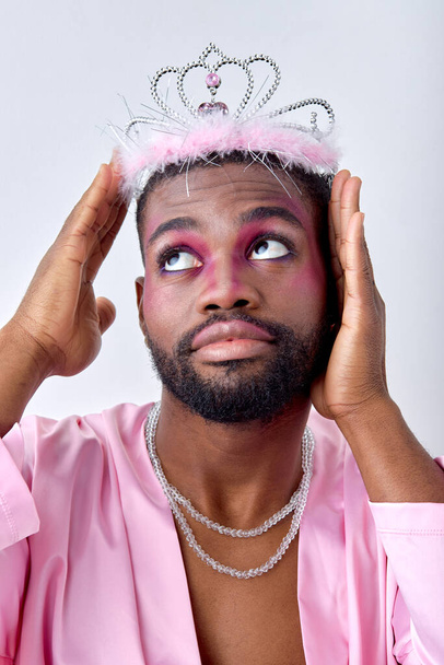 black guy in pink outfit, crown and necklace posing looking up, isolated over white studio background. Close-up portrait of feminine man with beard, american transgender straighten the crown - Фото, изображение