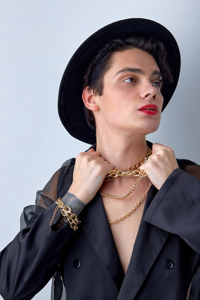 Attractive young transgender man LGBTQ being as woman posing with chain, in hat, gorgeous caucasian guy showing self-confidence, with red lipstick and make-up isolated on white background. - Foto, immagini